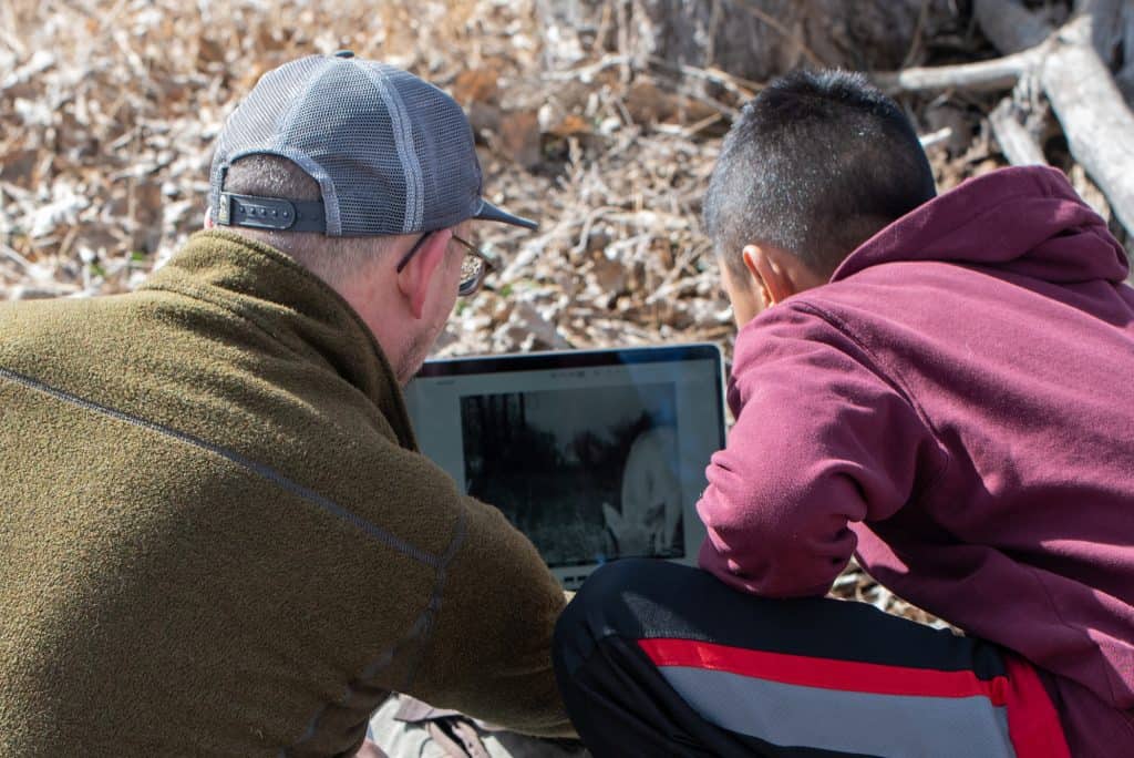 Looking from behind, a male teacher on the left and a male middle school student on the right are sitting on the ground in an open space area looking at photos from the SD card they removed from their trail camera. A deer can be seen on the screen of the laptop.