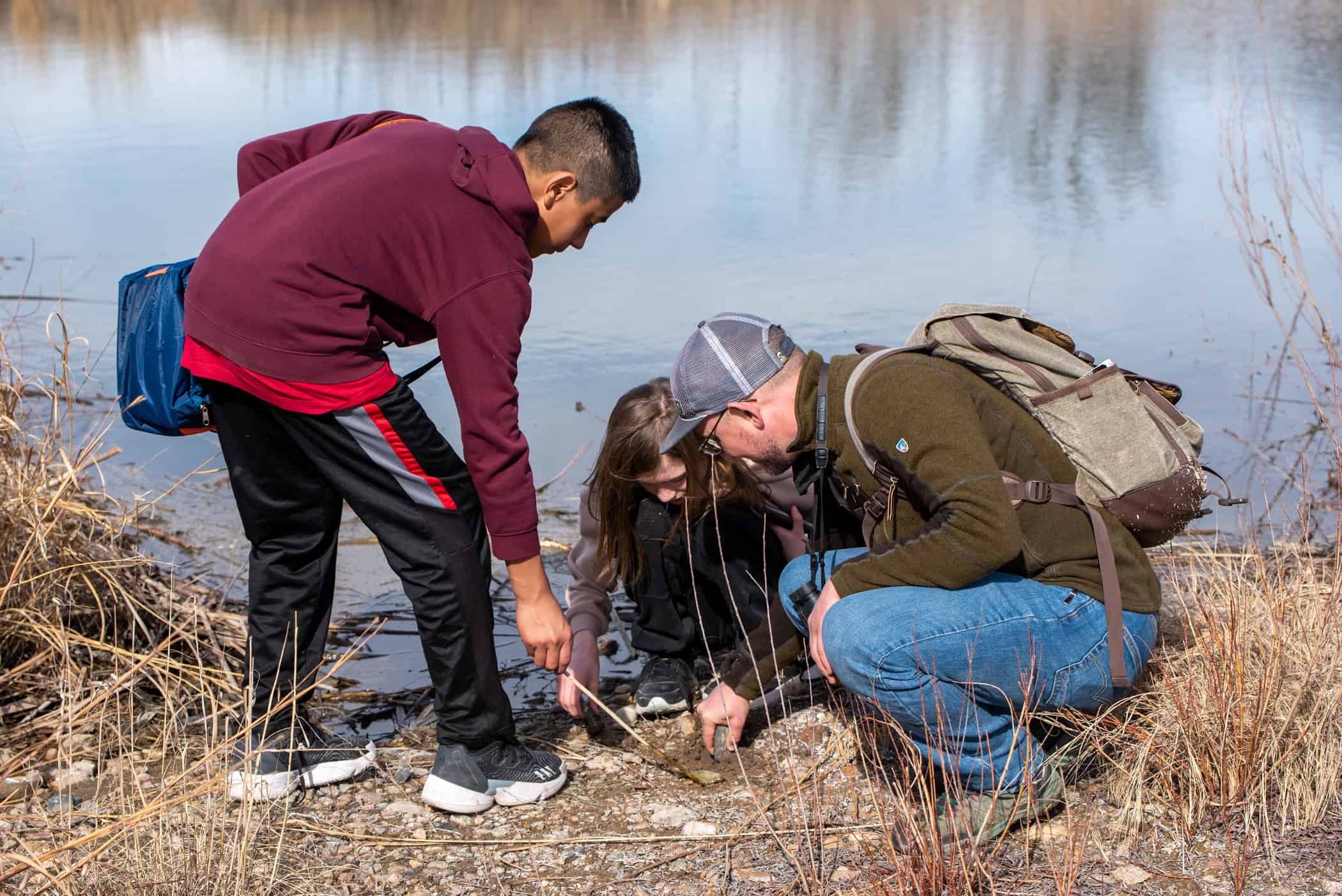 Male middle school student is bending over while pushing a stick into the ground next to a stream. A female middle school student and male teacher are squatting down examining something in the dirt to the right of the male student.