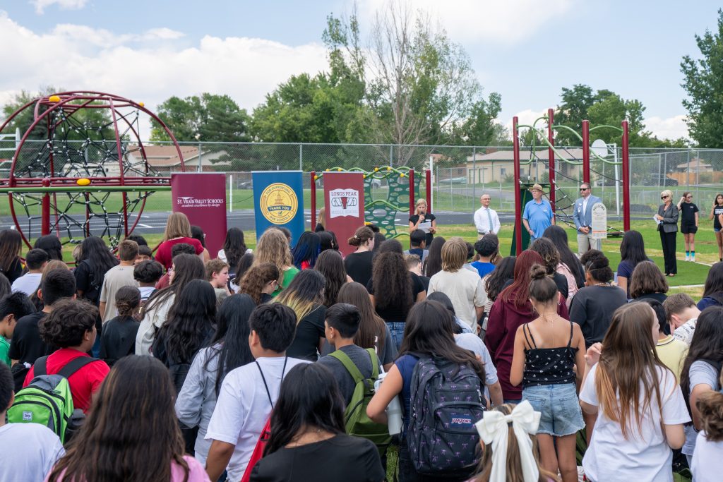 A crowd of students facing a stage with speakers, near a playground on a sunny day. Banners are visible.