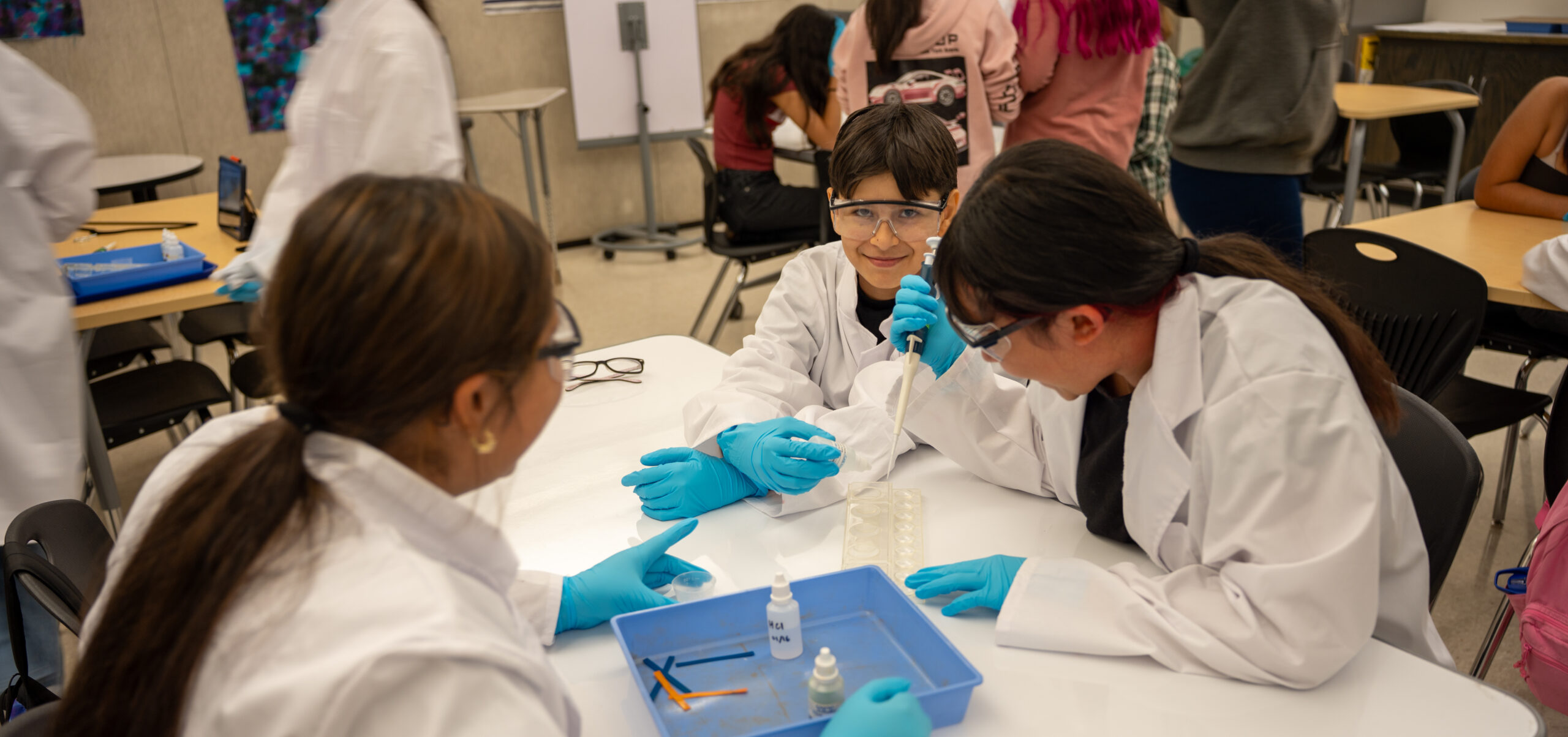 Students in lab coats conduct an experiment at a table, using pipettes and test samples in a classroom setting.