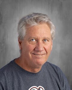 Smiling middle-aged man with light gray hair, wearing a dark t-shirt, against a neutral gray background.