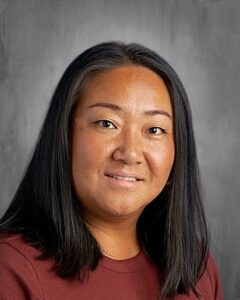 A woman with long black hair and a warm smile is wearing a maroon shirt against a gray background.