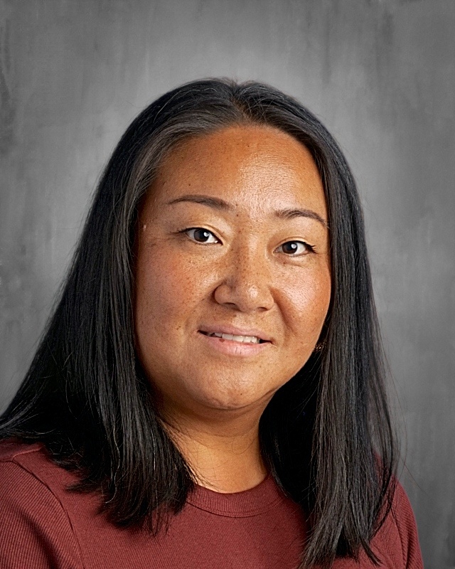 A woman with long black hair and a warm smile is wearing a maroon shirt against a gray background.