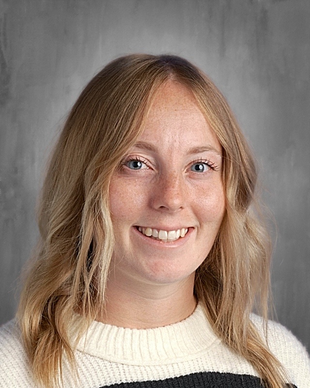 Smiling woman with long, wavy blonde hair, wearing a white and black sweater, against a gray background.