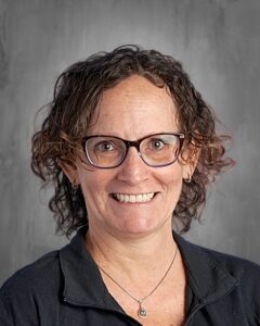 Mujer sonriente con el pelo rizado, gafas y una chaqueta negra, sobre un fondo gris.
