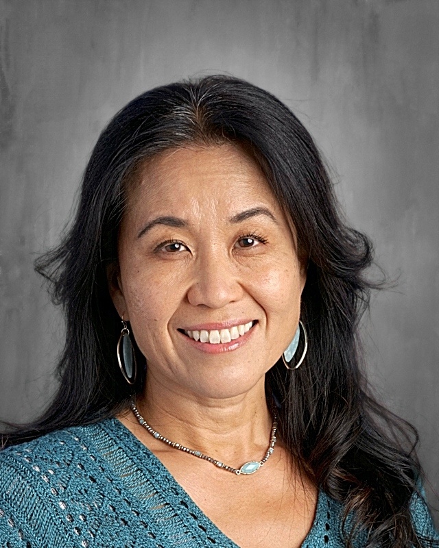 A smiling woman with long black hair, wearing a teal sweater and earrings, poses against a neutral background.