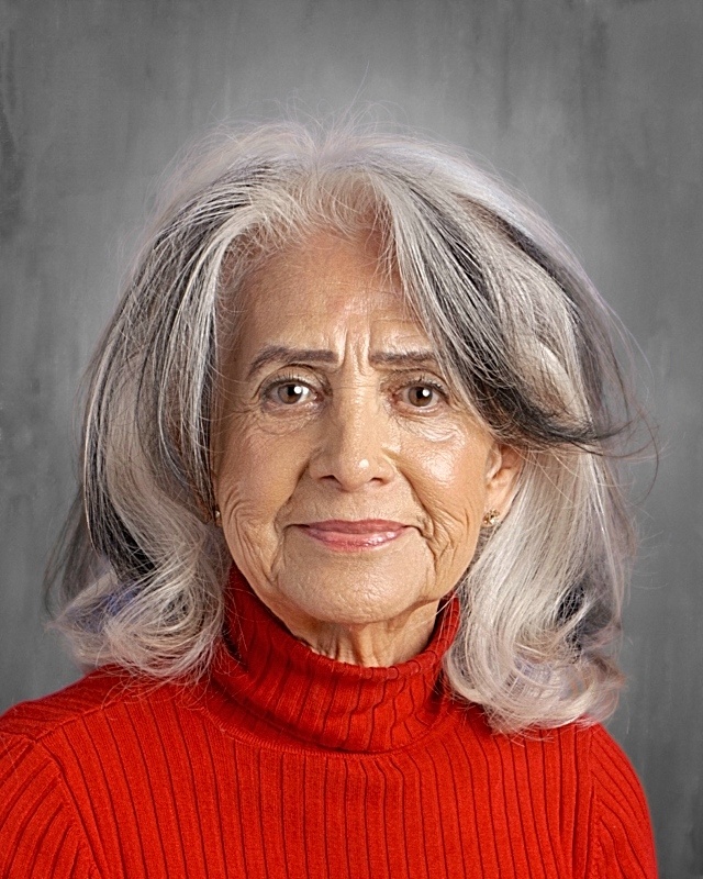 Senior woman with long, wavy gray hair wearing a red turtleneck sweater, smiling against a neutral background.
