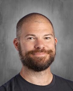 Smiling man with a short beard, wearing a black t-shirt, against a gray background.
