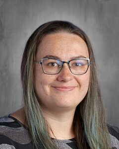 A woman with long hair and glasses smiles at the camera, wearing a patterned top against a gray background.