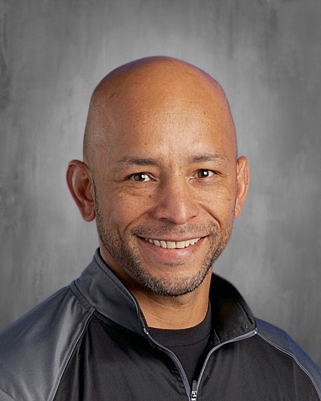 Smiling bald man in a black jacket, against a gray background. He has a light beard and friendly expression.