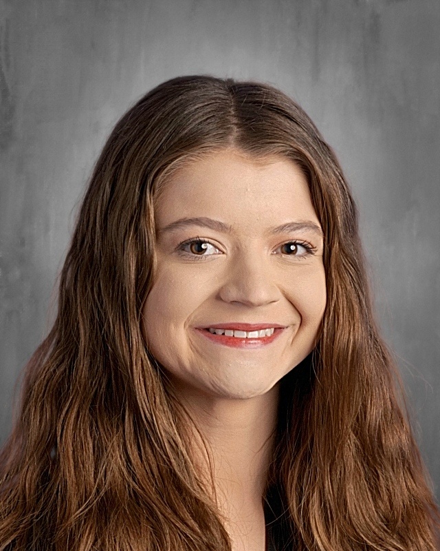 A smiling young woman with long, wavy brown hair, wearing a black top, against a gray background.