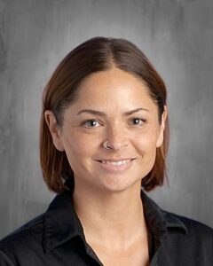 Portrait of a woman with short brown hair and a nose ring, smiling against a textured gray background.