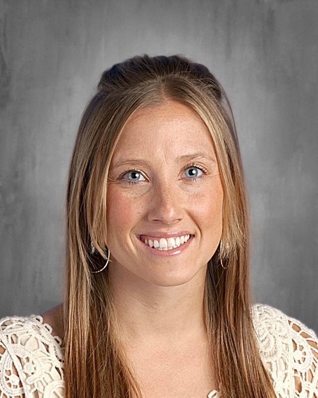 Smiling woman with long brown hair and blue eyes, wearing a light-colored top, set against a gray background.