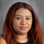 Close-up portrait of a woman with long, wavy brown hair, wearing a black top, against a gray background.