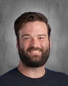Smiling man with a beard, wearing a dark t-shirt, against a gray background.