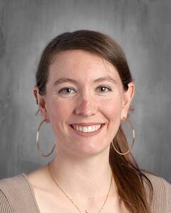A smiling woman with long brown hair, wearing hoop earrings and a beige top, against a gray background.