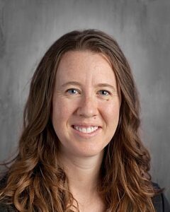 Smiling woman with long, wavy brown hair and blue eyes, set against a gray background.