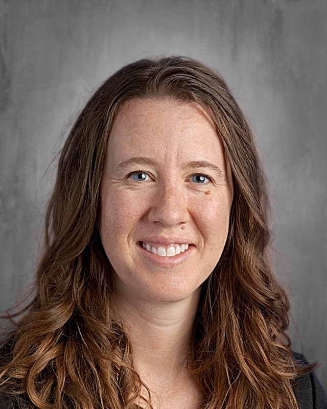 Smiling woman with long, wavy brown hair and blue eyes, set against a gray background.