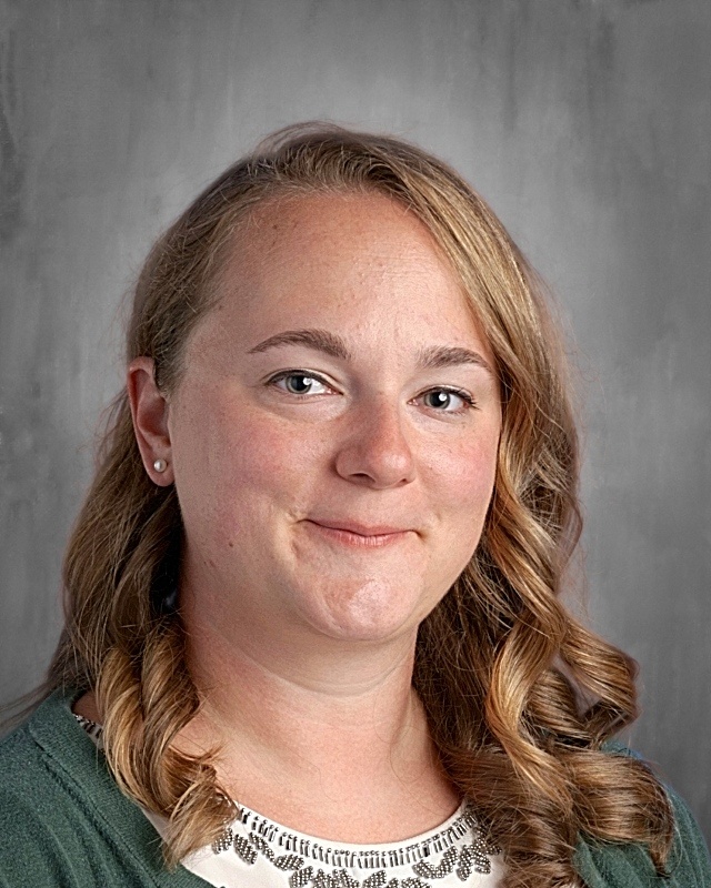 A woman with curly hair smiles for the camera, wearing a green sweater and a decorated top, against a gray background.