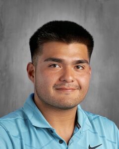 Young man with short black hair, smiling, wearing a light blue polo shirt against a gray background.