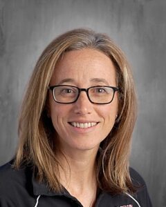 A smiling woman with glasses and shoulder-length hair, wearing a black shirt, poses against a gray background.