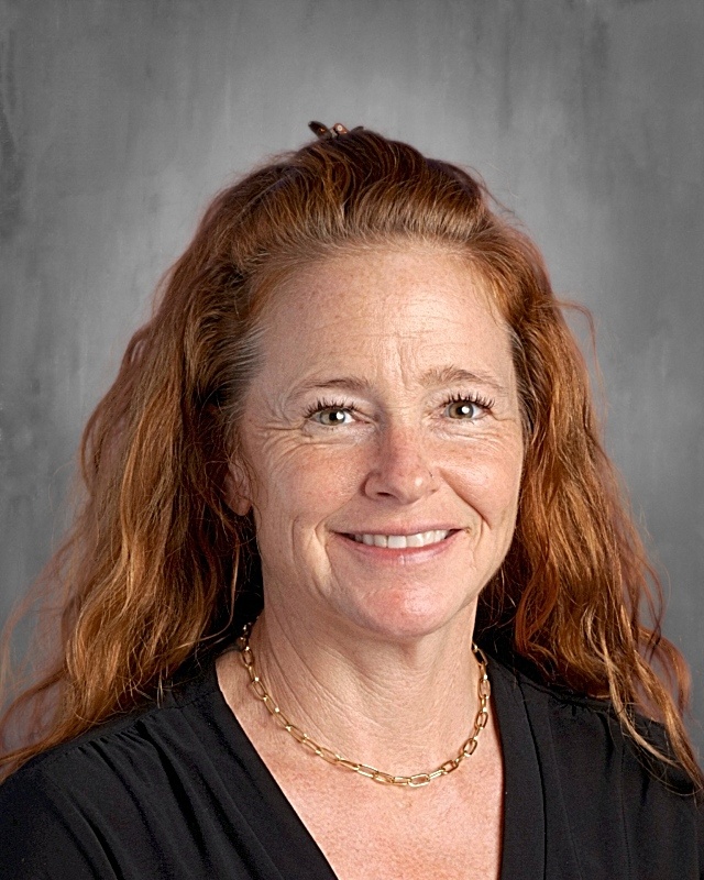 A woman with long, wavy red hair smiles, wearing a black top and a gold necklace, against a gray background.