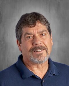 Smiling man with short gray hair and a beard, wearing a blue polo shirt, against a gray background.