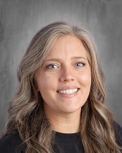 Headshot of a woman with long, wavy gray hair and blue eyes, smiling against a neutral gray background.