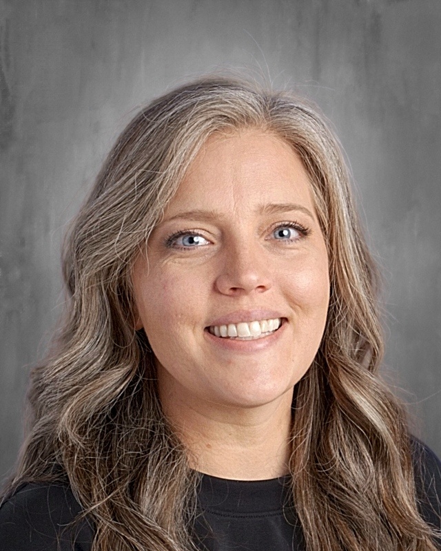 Headshot of a woman with long, wavy gray hair and blue eyes, smiling against a neutral gray background.
