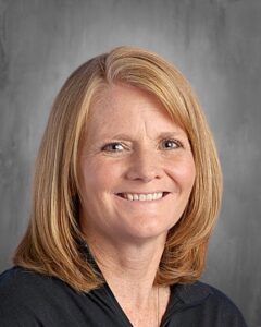 Smiling woman with shoulder-length blonde hair, wearing a black top, against a gray background.