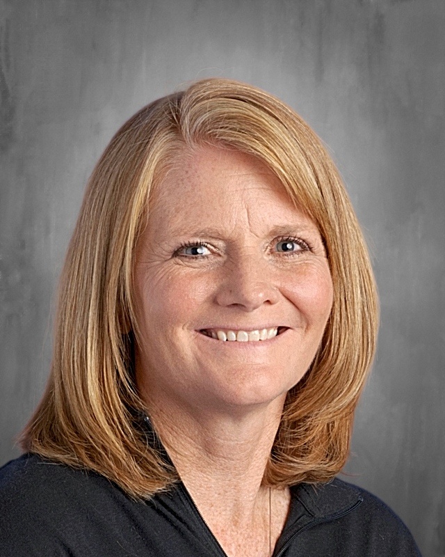 Smiling woman with shoulder-length blonde hair, wearing a black top, against a gray background.